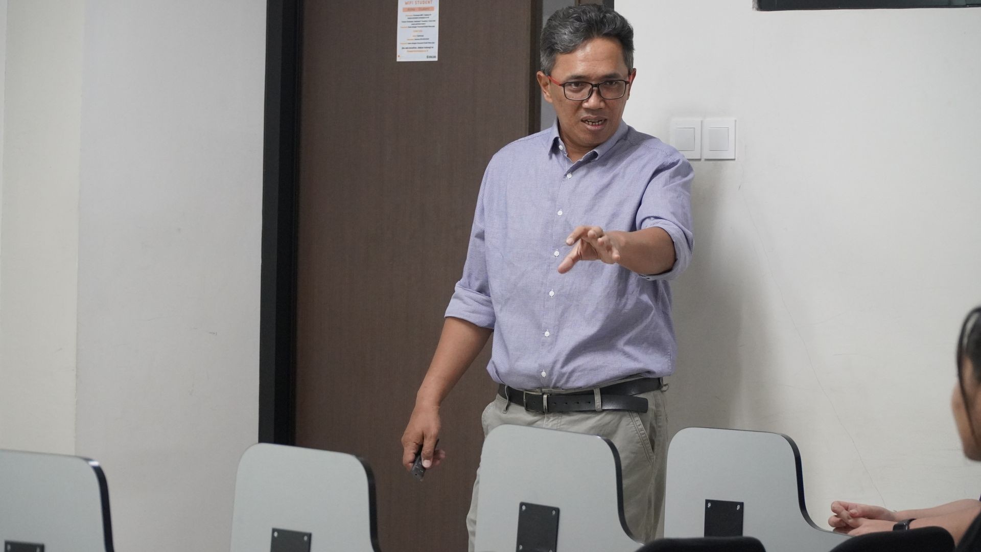Person standing and gesturing in front of a classroom with a brown door and white walls.