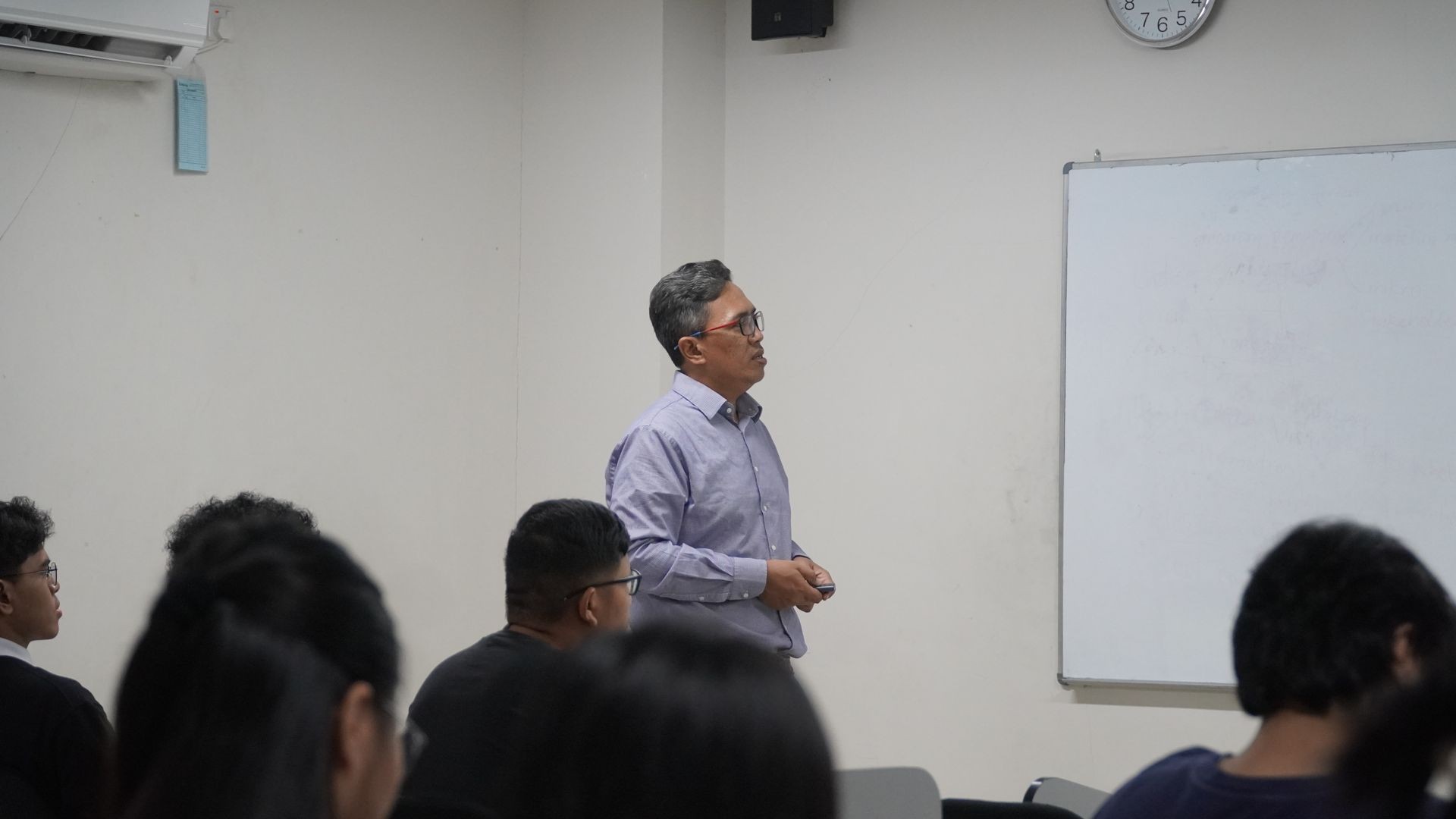 Person standing in a classroom setting next to a whiteboard, with students seated and facing forward.